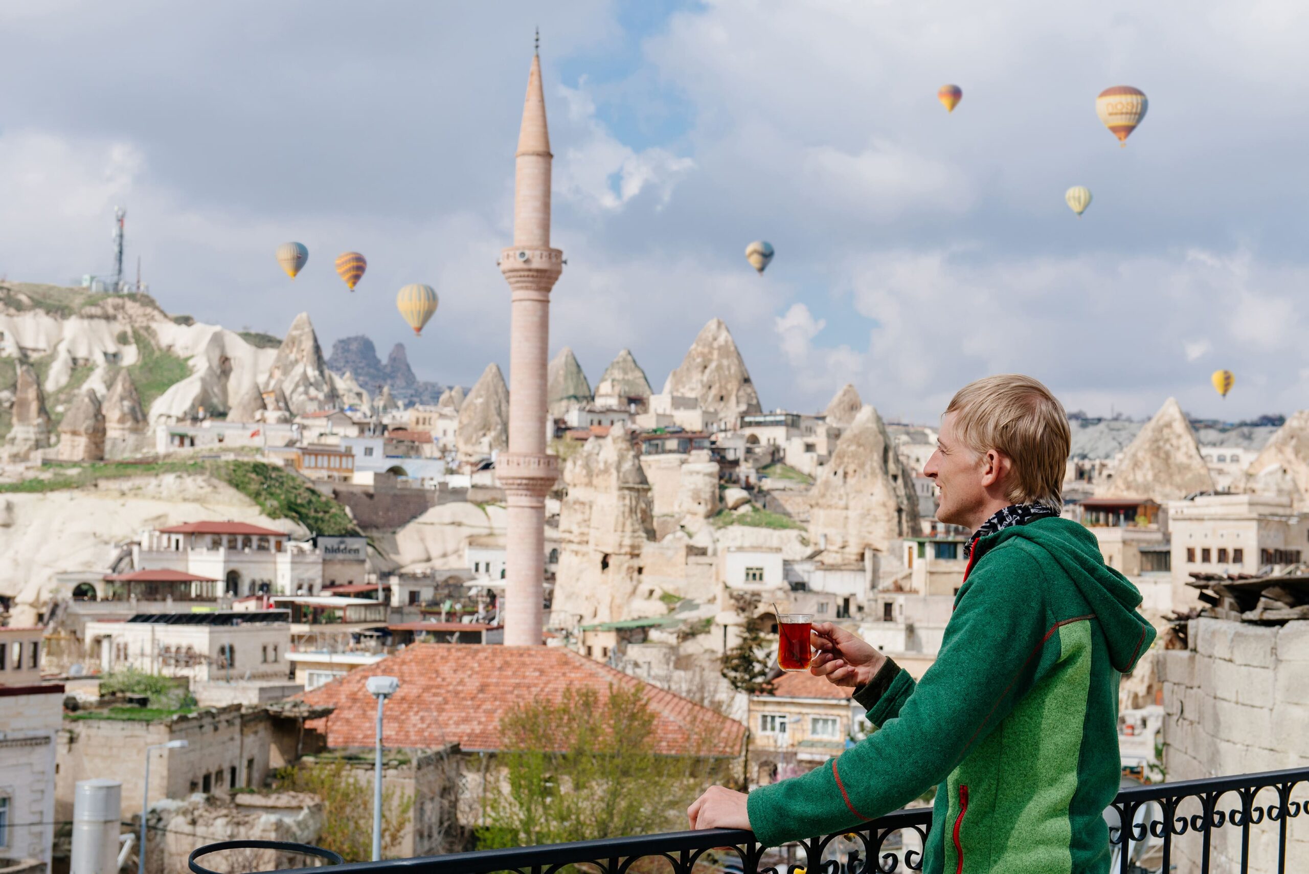 Man drinking turkish tea standing in front of old city Goreme, Cappadocia looking at air ballon in bring sunny day
